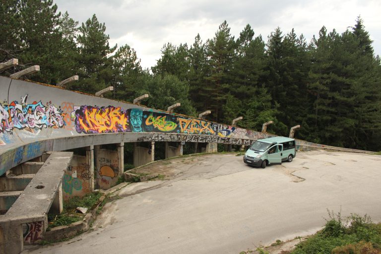 A light blue camper van is parked on a concrete road beside a curved, derelict structure covered in colourful graffiti, surrounded by tall green pine trees under a cloudy sky.